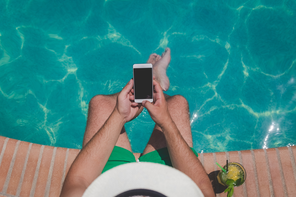 Un homme les pieds dans l'eau de sa piscine avec son téléphone à la main et un cocktail