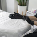 A cosmetologist is getting ready to perform a skincare treatment by putting on black gloves. The salon has a clean and professional atmosphere with elegant decor